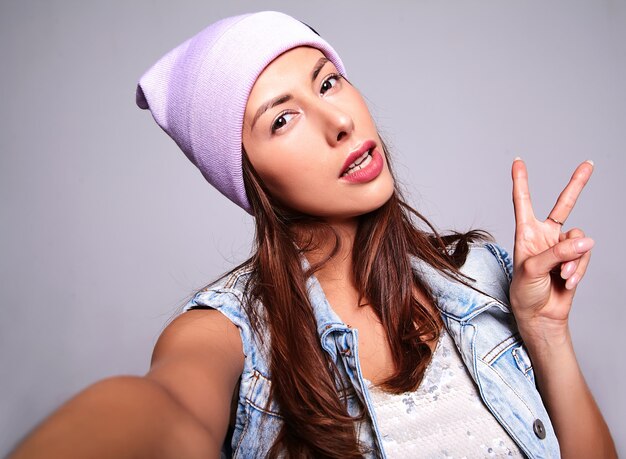 グレーに分離された携帯電話でselfie写真を作る紫色のビーニーでメイクなしのカジュアルな夏のジーンズ服で美しいかわいいブルネットの女性モデルの肖像画。ピースサインを表示