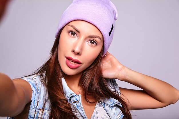 Portrait of beautiful cute brunette woman model in casual summer jeans clothes with no makeup in purple beanie making selfie photo on phone isolated on gray