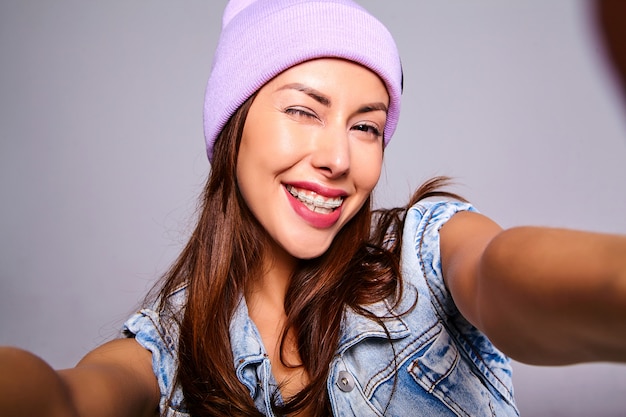 Il ritratto di bello modello sveglio della donna castana in jeans casuali dell'estate copre senza trucco in berretto porpora che fa la foto del selfie sul telefono isolato su gray