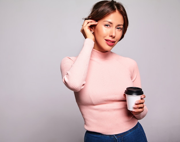 Portrait of beautiful cute brunette woman model in casual summer pink clothes with no makeup isolated on gray. Drinking coffee