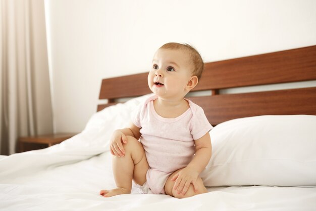Portrait of beautiful cute nice newborn baby in pink shirt sitting on bed at home.