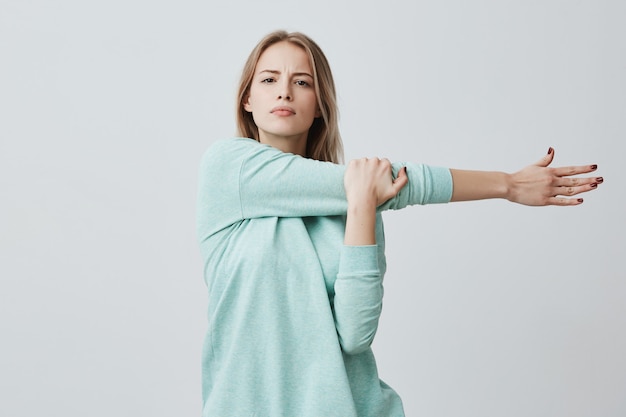 Free photo portrait of beautiful european woman with blonde long hair wearing casual blue sweater stretching her arm, doing exercises, cares about her health. fitness, health and beauty concept.