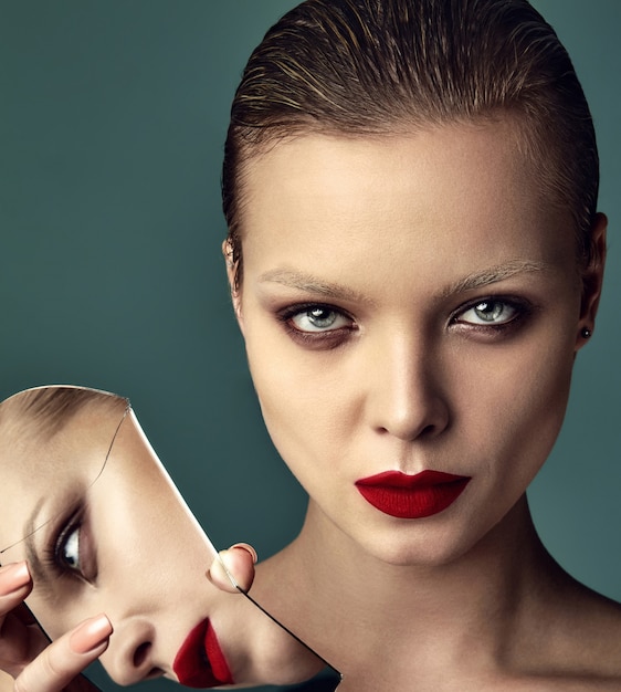 Portrait of beautiful fashion stylish brunette woman model with evening makeup and red lips reflecting in broken mirror on blue