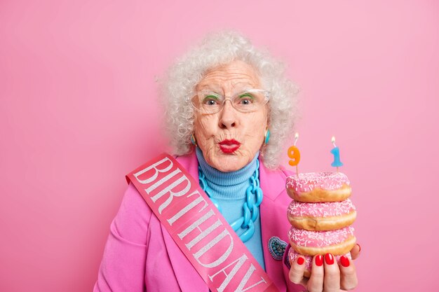 Portrait of beautiful female pensioner keeps lips rounded wants to kiss you and says thankful for congratulations being well dressed holds pile of delicious doughnuts