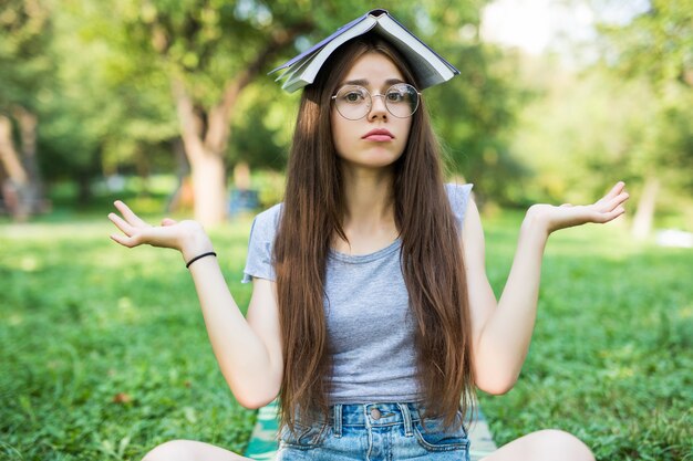 그녀의 머리 위에 카피 북 앨범을 들고 안경을 쓰고 푸른 잔디에 야외 공원에 앉아 아름 다운 재미 귀여운 행복 젊은 아가씨 여자 학생의 초상화.