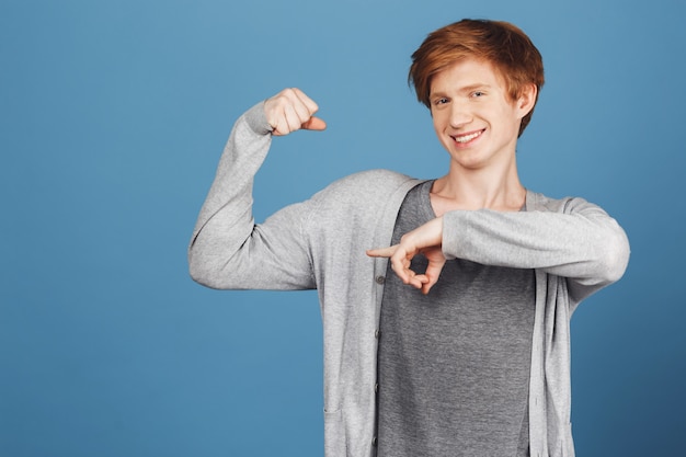 Free photo portrait of beautiful ginger young guy on blue wall wearing casual grey outfit pointing with index finger on his muscles,  with happy and satisfied expression.