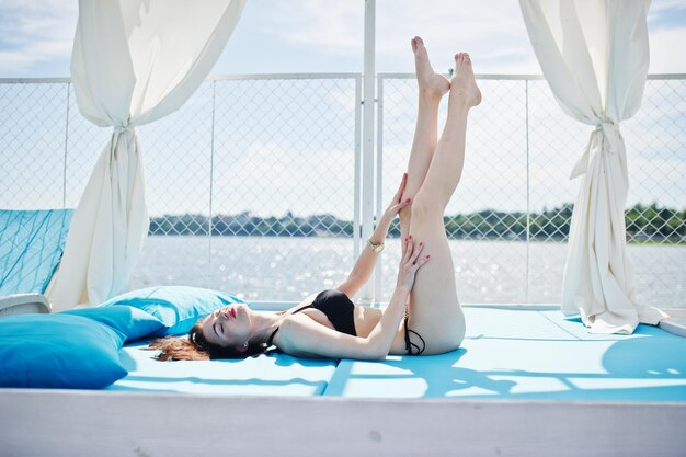 Portrait of a beautiful girl in black bikini swimsuit laying on the lounger by the lakeside