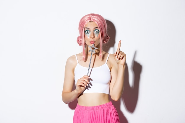 Portrait of beautiful girl in fairy costume looking thoughtful, holding magic wand and wearing pink wig, have an idea, standing.