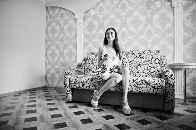 Портрет красивой девушки в цветочном платье, сидящей на диване с пультом в руках Черно-белое фото