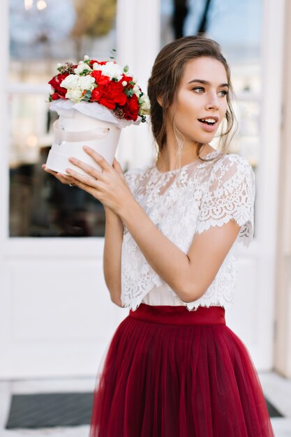 Portrait beautiful girl in marsala tulle skirt on street. She holds bouuquet of roses, looking to side