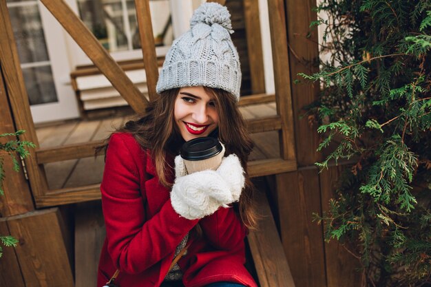 Портрет красивой девушки в красном пальто, вязаной шляпе и белых перчатках, сидя на деревянной лестнице на открытом воздухе. Она держит кофе и улыбается в сторону.