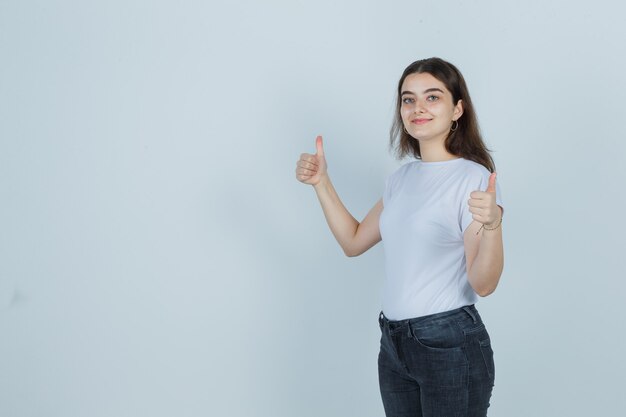 Portrait of beautiful girl showing thumbs up in t-shirt, jeans and looking confident, front view