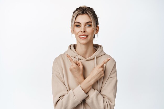 Portrait of beautiful girl smiles, shows two ways, points sideways left and right, choice gesture, stands in beige hoodie on white.