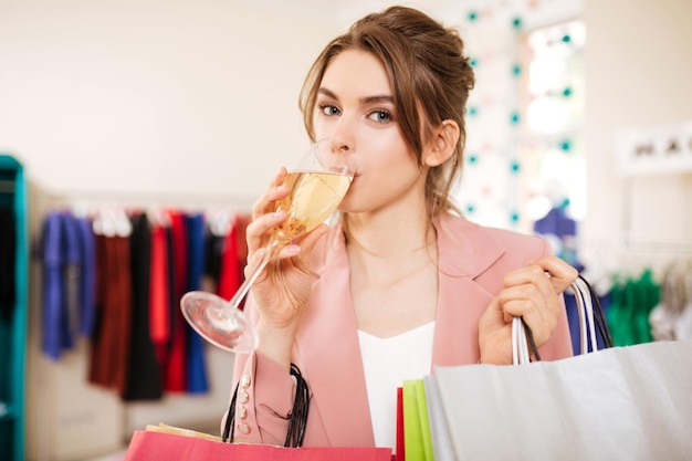 Free photo portrait of beautiful girl with brown hair in pink pantsuit drinking champagne and thoughtfully looking in camera while standing with shopping bags in fashion boutique