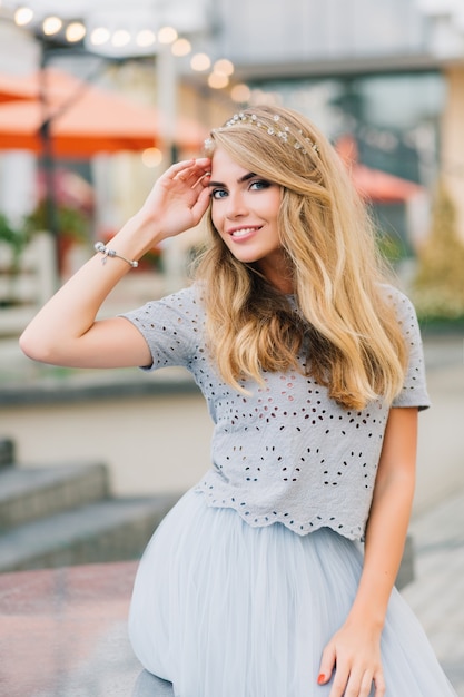 Portrait beautiful girl with long blonde hair in blue tulle skirt sitting on terrace background. She keeps hand on head and smiling to camera.