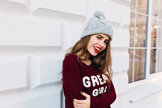세로 외부 회색 벽에 마르 살라 스웨터에 긴 머리를 가진 아름 다운 소녀. 그녀는 니트 모자, 붉은 입술을 쓰고 웃고 있습니다.