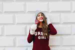 Free photo portrait beautiful girl with long hair  in marsala sweater and white gloves on grey wall . she touches knitted hat, holds red heart candy and smiling .