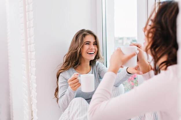 Portrait beautiful girl with long hair in pajama drinking tea on window in light room. She is smiling to girl in front