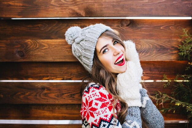 Portrait beautiful girl with long hair and red lips in knitted hat and gloves on wooden . She is smiling .