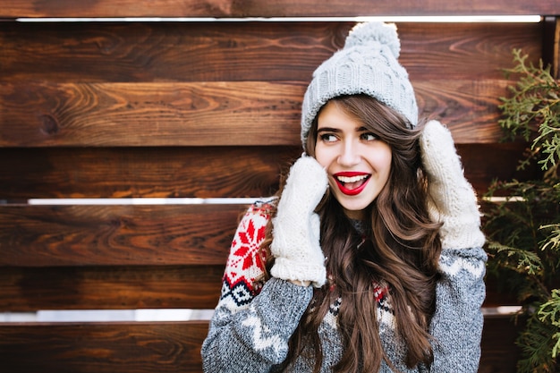 Portrait beautiful girl with long hair and red lips in knitted hat and warm gloves on wooden . She smiling to side.