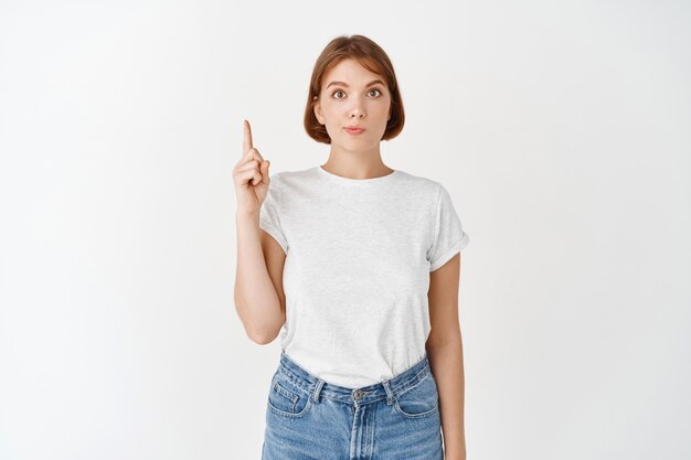 Portrait of beautiful girl with natural make-up, pointing finger up. Young woman student in casual clothes showing way on top, standing on white wall