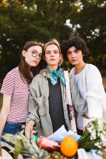 Portrait of beautiful girls with bicycle and basket full of wildflowers and fruits dreamily looking in camera spending time together in park