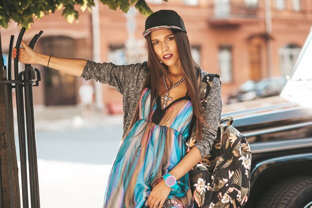 Portrait of beautiful glamor brunette teenager model in summer hipster clothes and bag. Girl posing in the street. Woman in cap