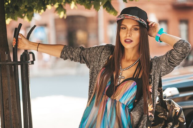 Portrait of beautiful glamor brunette teenager model in summer hipster clothes and bag. Girl posing in the street. Woman in cap