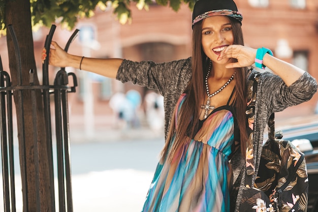 Portrait of beautiful glamor smiling brunette teenager model in summer hipster clothes and bag. Girl posing in the street. Woman in cap