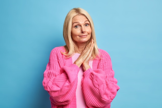 Portrait of beautiful grandma keeps palms pressed together and looks curiously dressed in knitted rosy jumper enjoys life on retirement.