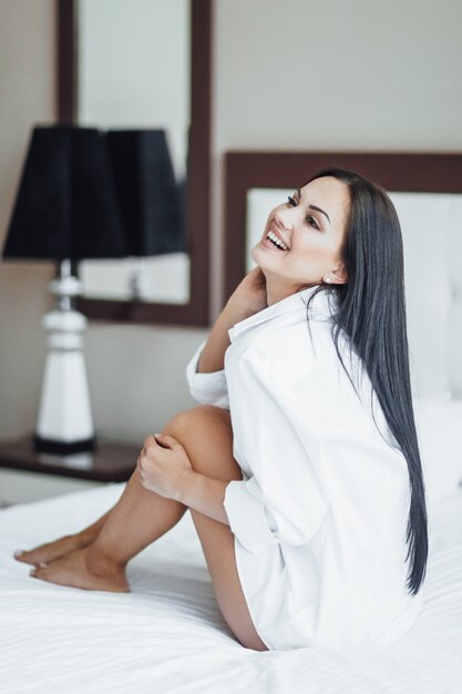 Портрет красивой счастливой девушки брюнет сидя в кровати и позируя.
