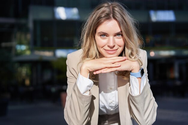 Portrait of beautiful happy corporate woman in suit stands on street and smiles poses near office bu