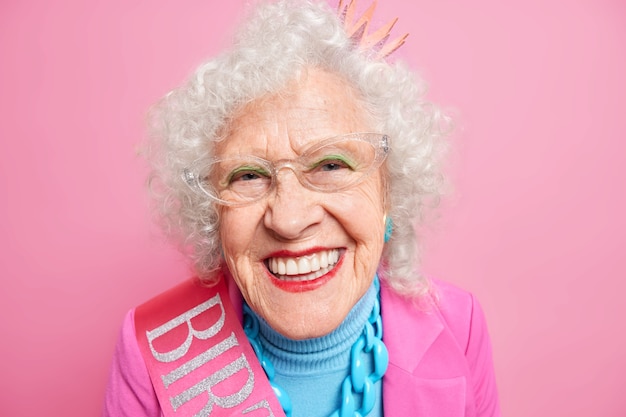 Portrait of beautiful happy grandmother smiles toothily wears red lipstick has white perfect teeth dressed in festive clothes enjoys retirement expresses positive emotions. People age beauty concept