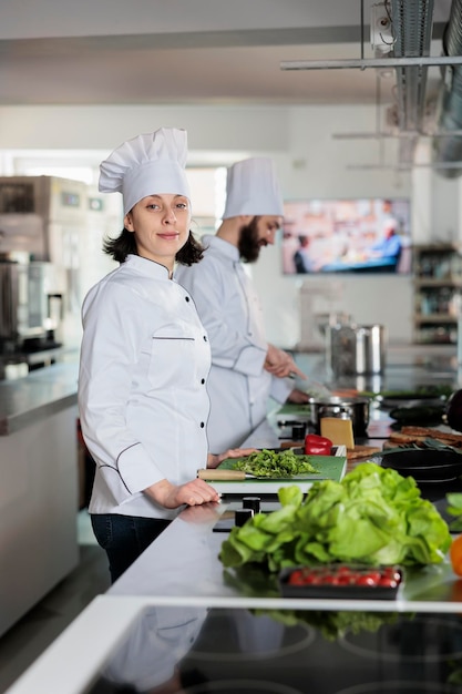 Portrait of beautiful happy sous chef standing in restaurant professional kitchen, wearing cooking uniform while smiling at camera. Young adult food industry worker preparing vegetables for meal.