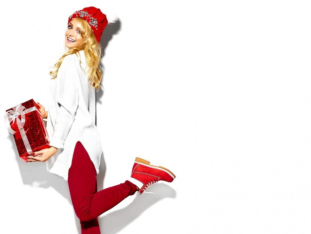 portrait of beautiful happy sweet smiling blonde woman girl holding in her hands big Christmas gift box in casual red hipster winter clothes, in white warm sweater standing on one leg