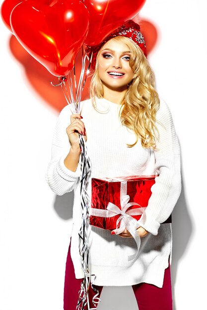 portrait of beautiful happy sweet smiling blonde woman girl holding in her hands big Christmas gift box and heart balloons in casual red hipster winter clothes, in white warm sweater