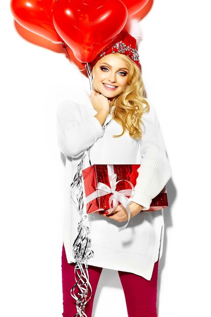 portrait of beautiful happy sweet smiling blonde woman girl holding in her hands big Christmas gift box and heart balloons in casual red hipster winter clothes, in white warm sweater