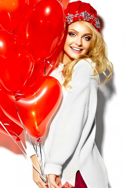 portrait of beautiful happy sweet smiling blonde woman girl holding in her hands big Christmas gift box and heart balloons in casual red hipster winter clothes, in white warm sweater