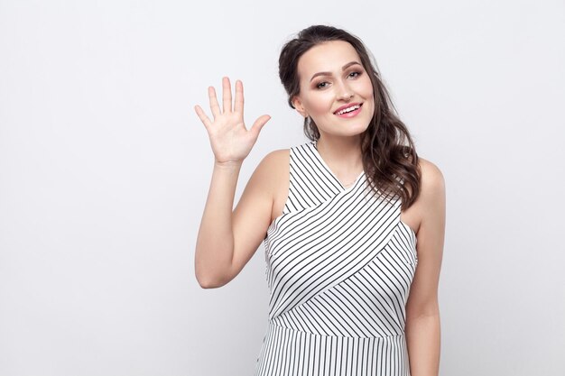 Portrait of beautiful happy young brunette woman with makeup and striped dress standing, looking at camera with toothy smile and greeting gesture. indoor studio shot, isolated on grey background.