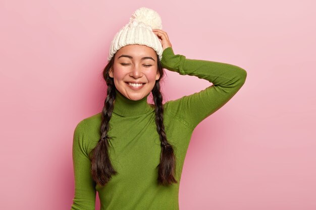 Portrait of beautiful hipster girl, keeps eyes shut, dressed in green poloneck and hat, satisfied after spending free time with close person remembers pleasant moment during romantic date