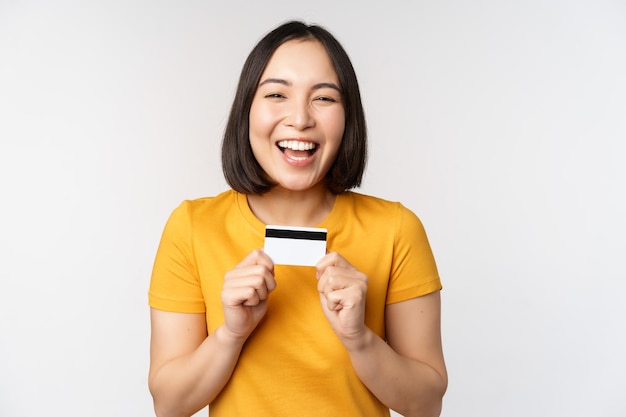 白い背景の上に黄色のTシャツで立っている銀行サービスをお勧めするクレジットカードを保持している美しい韓国の女の子の肖像画