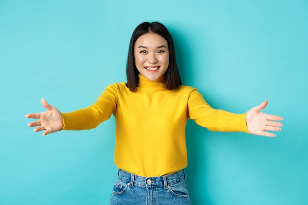 Free photo portrait of beautiful korean woman spread out hands for hug, reaching for cuddles and smiling at camera, greeting you, standing over blue background.