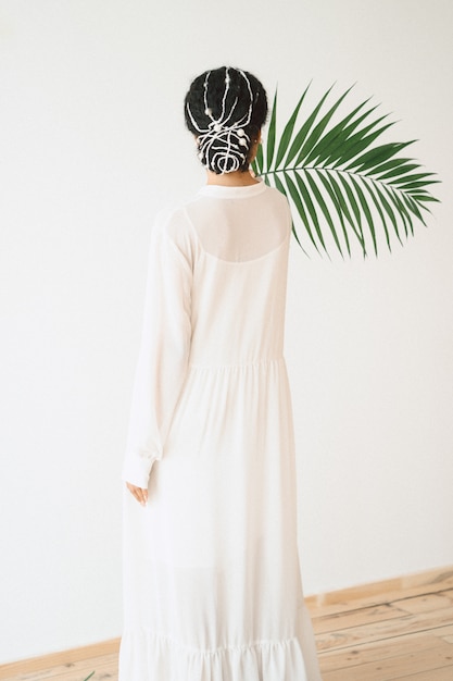 Free photo portrait of beautiful lady at empty room standing and holding palm branch in long white dress