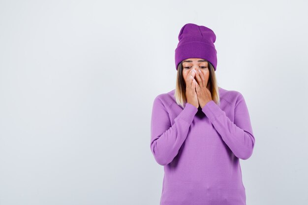 Portrait of beautiful lady keeping hands on face in sweater, beanie and looking shocked front view