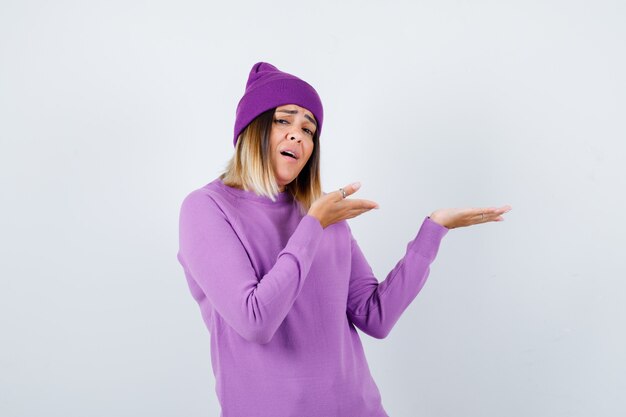 Portrait of beautiful lady pretending to show something in sweater, beanie and looking troubled front view