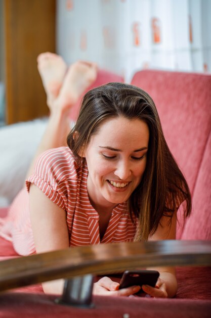 Free photo portrait of beautiful lady at room on a couch lying, looking at her phone and smiling in pink striped dress