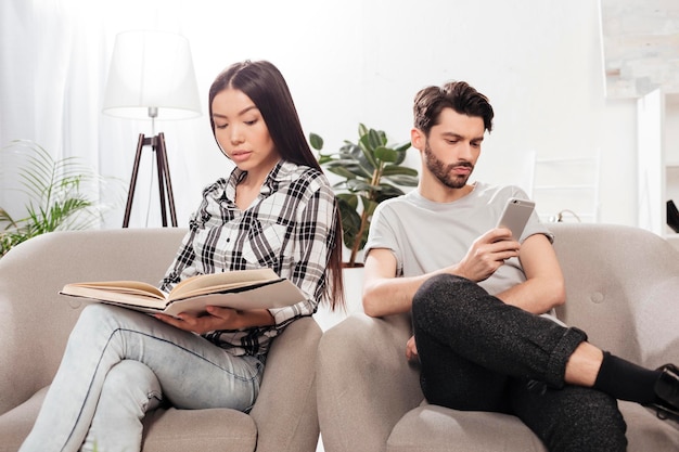 Portrait of beautiful lady sitting in chair and reading book with young man near sitting and using his cellphone at home isolated