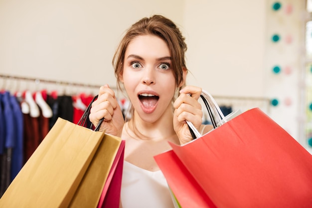 Ritratto di bella signora con i capelli castani in tailleur rosa che guarda stupito nella fotocamera mentre è in piedi con le borse della spesa in mano nella boutique di moda