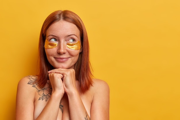 Portrait of beautiful middle aged woman keeps hands under chin, looks aside thoughtfully, enjoys soft smooth skin, wears beauty patches to reduce fine lines, isolated on yellow wall with blank space