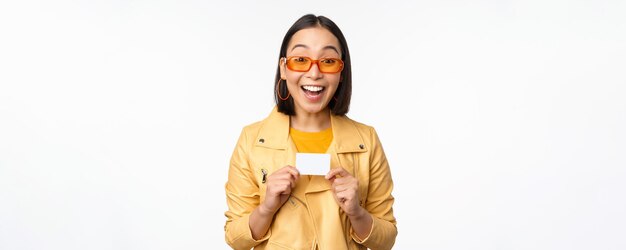 白い背景の上に立っているクレジットカードを示して幸せに笑ってサングラスで美しい現代アジアの女の子の肖像画